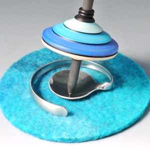 Könnte beinhalten: Ein Kreisel mit abgestuftem Design in Hellblau, Mittelblau und Dunkelblau sowie einer dunkelgrauen Mittelachse. Die Spitze des Kreisels ruht auf einer kreisförmigen, silberfarbenen Metallbasis mit einem geschwungenen, offenen Ring.