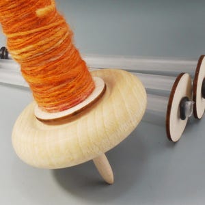May include: A wooden drop spindle with bright orange and yellow yarn. The spindle is made of light-coloured wood and has a rounded shape. The yarn is tightly wound around the spindle. Additional clear tubes and wooden discs are visible, for storing the yarn.