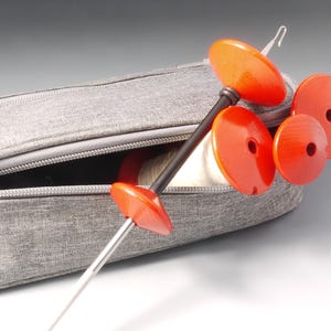 May include: A grey zipped case contains a set of orange and black knitting tools. The tools include a long, thin needle with an orange handle and several orange, disc-shaped components. The case is open, revealing the tools inside.
