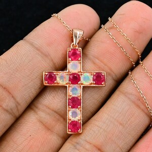May include: A gold-colored cross pendant necklace. The cross is adorned with alternating red and white gemstones. The necklace chain is also gold-colored. The pendant is held in a person's hand against a black background.
