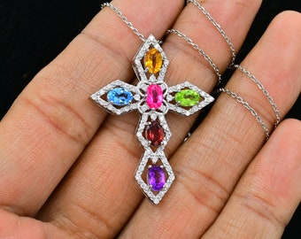 Puede incluir: Un collar con un colgante de cruz de plata con un diseño incrustado de diamantes blancos. La cruz presenta siete piedras preciosas ovaladas de varios colores, incluyendo azul, rosa, verde, naranja, rojo y morado.