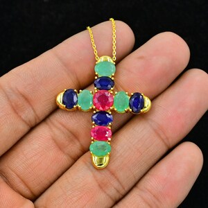Puede incluir: Collar con colgante de cruz de oro con piedras preciosas ovaladas en tonos de verde, rojo y azul.