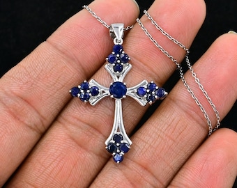 May include: A silver cross pendant with blue sapphire gemstones. The cross is detailed with intricate designs and features a round sapphire in the center.