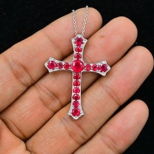 May include: A silver cross pendant with red ruby gemstones. The cross is detailed with intricate designs and is set with round rubies.