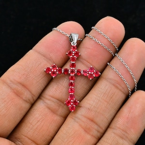 Puede incluir: Un colgante de cruz de plata con piedras preciosas rojas. La cruz está engastada con pequeñas piedras redondas rojas que cubren toda la superficie. El colgante está unido a una cadena de plata.
