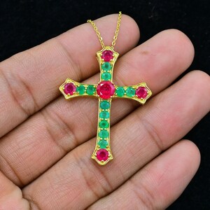 Puede incluir: Un colgante de cruz de oro con piedras preciosas rojas y verdes. La cruz está engastada con piedras redondas y facetadas. El colgante está en una cadena de oro.