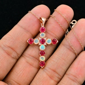 Puede incluir: Un collar con un colgante de cruz de oro rosa con rubíes rojos y opales blancos. La cruz está adornada con pequeños diamantes transparentes.