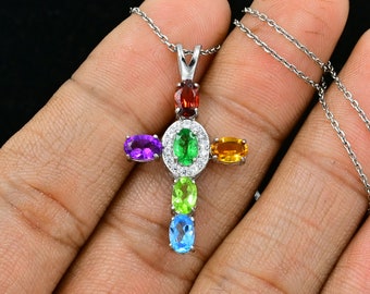 Puede incluir: Un collar de cadena de plata con un colgante de cruz que presenta siete piedras preciosas ovaladas en colores arcoíris. Las piedras preciosas están engastadas en un marco de plata con un halo de piedras transparentes más pequeñas.