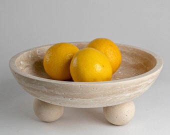 May include: A light beige travertine bowl with three legs and three yellow lemons inside. The bowl is on a white surface.