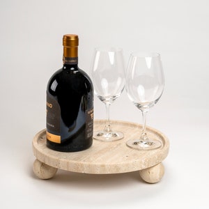 May include: A round, light brown, travertine serving tray with three legs. The tray has a bottle of red wine and two clear wine glasses on top.
