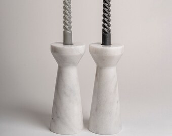 Op de afbeelding: Twee witte marmeren kandelaars met een uitlopende voet en een brede opening aan de bovenkant. Elke kandelaar bevat een lange, slanke, gedraaide taps toelopende kaars. De linkerkaars is lichtgrijs en de rechterkaars is donker antraciet, beide brandend.