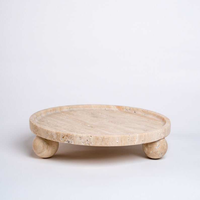 May include: A round, beige travertine tray with a smooth surface and three rounded legs. The tray is made of natural stone and has a rustic, organic look.