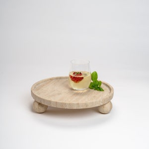 May include: A round, light beige, travertine serving tray with three rounded legs. A clear glass with a strawberry inside and a white liquid is on the tray. A sprig of green basil is next to the glass.