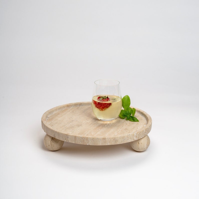 May include: A round, light beige, travertine serving tray with three rounded legs. A clear glass with a strawberry inside and a white liquid is on the tray. A sprig of green basil is next to the glass.