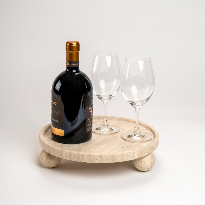 May include: A round, light brown, travertine serving tray with three legs. The tray has a bottle of red wine and two clear wine glasses on top.