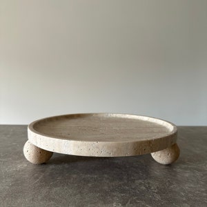 May include: A round, off-white, stone tray with three round legs. The tray is made of travertine and has a rough, textured surface.