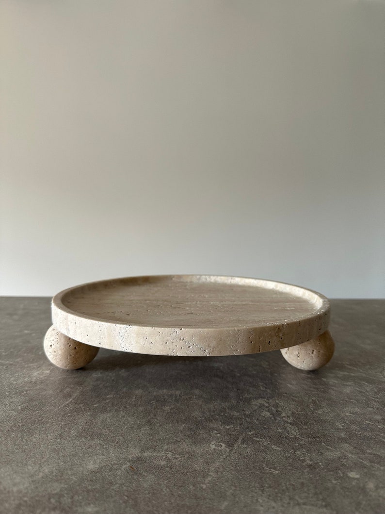 May include: A round, off-white, stone tray with three round legs. The tray is made of travertine and has a rough, textured surface.