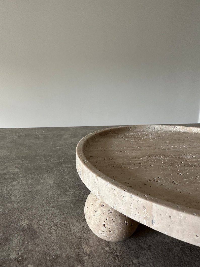 May include: A round, light beige stone tray with a raised edge. The tray is supported by three round, light beige stone balls.