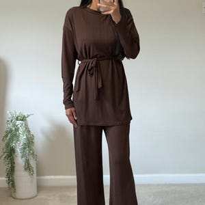 Op de afbeelding: Een bruin geribbelde gebreide tweedelige outfit. De top met lange mouwen heeft een strikceintuur in de taille. De outfit bevat een broek met wijde pijpen. Het model draagt gouden oorringen.