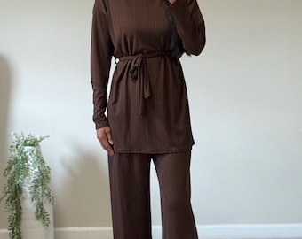 May include: A brown ribbed knit two-piece outfit. The long-sleeved top has a tie belt at the waist. The outfit includes wide-leg pants. The model is wearing gold hoop earrings.