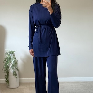 May include: Navy blue two-piece outfit. The set includes a long-sleeved top with a tie waist and wide-leg pants. The outfit is made of a ribbed fabric. The person in the image is wearing gold-colored sandals and earrings.