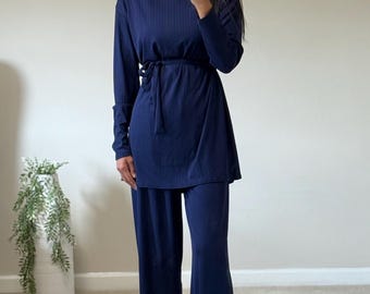 May include: Navy blue two-piece outfit. The set includes a long-sleeved top with a tie waist and wide-leg pants. The outfit is made of a ribbed fabric. The person in the image is wearing gold-colored sandals and earrings.