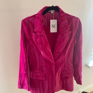 May include: A pink velvet blazer with a single button closure and a pocket on the left side. The blazer has a textured, slightly wrinkled appearance. The label on the blazer reads "NK."