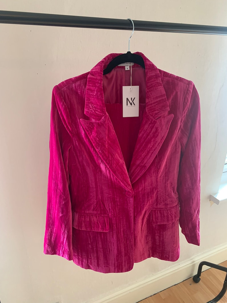 May include: A pink velvet blazer with a single button closure and a pocket on the left side. The blazer has a textured, slightly wrinkled appearance. The label on the blazer reads "NK."