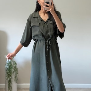 May include: Olive green button-down dress with short sleeves and a tie waist. The dress features a collared neckline, two chest pockets, and a long, flowing skirt. The dress is made of a lightweight fabric and has a relaxed fit, suitable for casual wear.