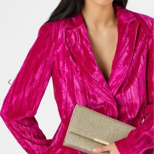 May include: A gold glitter clutch purse with a gold clasp. The purse is held by a person wearing a pink velvet blazer.