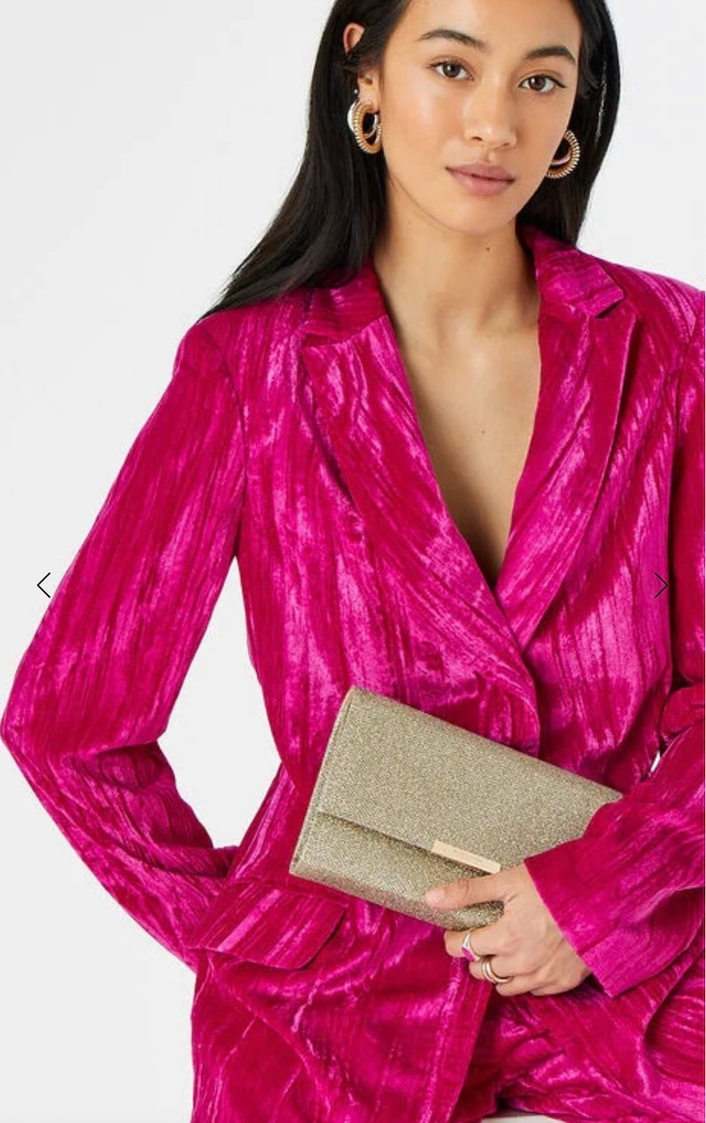 May include: A gold glitter clutch purse with a gold clasp. The purse is held by a person wearing a pink velvet blazer.