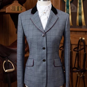 May include: A grey equestrian show jacket featuring a dark windowpane check pattern, a black velvet collar, and thin maroon piping along the lapels and pocket flaps. The jacket has three front buttons and is worn over a white stock tie with a pearl pin.