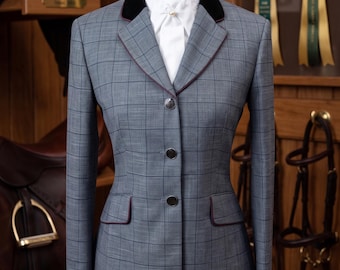 May include: A grey equestrian show jacket featuring a dark windowpane check pattern, a black velvet collar, and thin maroon piping along the lapels and pocket flaps. The jacket has three front buttons and is worn over a white stock tie with a pearl pin.