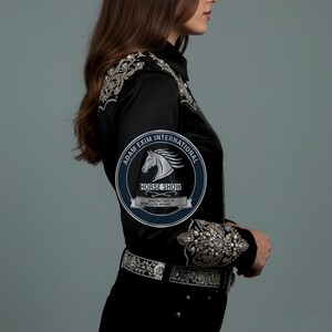 Op de afbeelding: Zwart shirt met lange mouwen met sierlijke zilveren en heldere kristallen versieringen op de schouders en manchetten. Het shirt heeft een kraag en een decoratieve riem. Het Adam Exim International Horse Show-logo is zichtbaar.