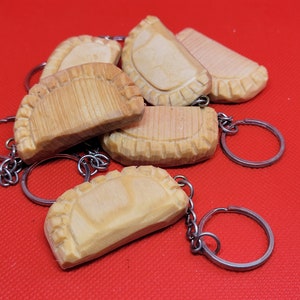 May include: A selection of wooden pierogi keyrings. Each keyring showcases a carved pierogi with detailed ridges and a light brown hue. The keyrings are attached to silver metal rings, set against a red backdrop.
