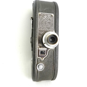 May include: A vintage Keystone 8mm movie camera, black with silver accents. The camera has a lens, dials, and a serial number plate. The text on the camera includes "Keystone" and exposure settings.