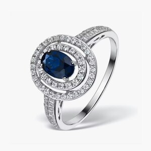 Könnte beinhalten: Ein silberner Ring mit einem großen ovalen blauen Saphir, der von einem doppelten Halo aus kleineren Diamanten umgeben ist.