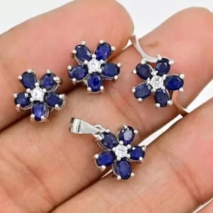 Könnte beinhalten: Ein silberner Blumen-Anhänger, Ohrringe und Ring-Set mit blauen und weißen Edelsteinen. Der Schmuck liegt auf einer Hand.