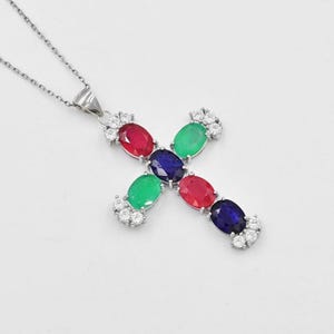 Puede incluir: Un collar con un colgante de cruz de plata con un diseño de piedras preciosas rojas, verdes, azules y blancas.