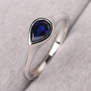 Puede incluir: Un anillo de compromiso de zafiro en forma de pera engastado en una banda de plata. El anillo tiene un diseño sencillo y presenta un solo zafiro grande.
