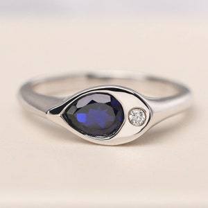 Könnte beinhalten: Ein silberner Ring mit einem tropfenförmigen, tiefblauen Edelstein und einem kleinen, runden, klaren Diamanten. Der Ring hat ein glattes, poliertes Band und eine augenähnliche Fassung. Der Ring ist ein Schmuckstück.