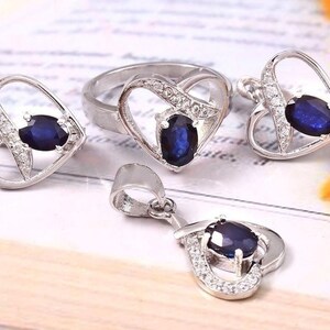 Könnte beinhalten: Ein silberner Herz-Ring, eine Halskette und Ohrringe mit einem blauen Saphir-Edelstein in jedem Stück. Der Schmuck ist mit kleinen, klaren Zirkonia-Steinen besetzt.