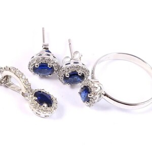 May include: A jewelry set featuring a silver pendant necklace, stud earrings, and a ring. Each piece has a deep blue oval gemstone surrounded by small, clear accent stones. The jewelry is set against a white background.