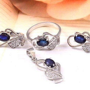 Könnte beinhalten: Ein silberner Ring, ein Paar Ohrringe und eine Halskette mit Anhänger, alle mit einem blauen ovalen Edelstein, der in einem herzförmigen Design mit weißen Akzenten gefasst ist.