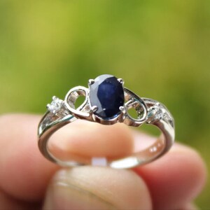 Könnte beinhalten: Ein silberner Ring mit einem großen ovalen blauen Saphir-Edelstein und zwei kleineren weißen Edelsteinen auf der Ringschiene.
