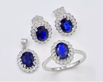May include: A jewellery set comprising a ring, pendant, and stud earrings. Each item showcases a large, oval, deep blue gemstone encircled by small, clear stones. The ring and earrings have a silver-coloured metal setting, and the pendant has a silver bail.