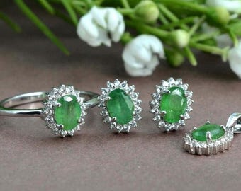May include: A set of emerald green gemstone jewelry featuring a ring, earrings, and a pendant. Each piece is set in silver with a halo of smaller white gemstones.