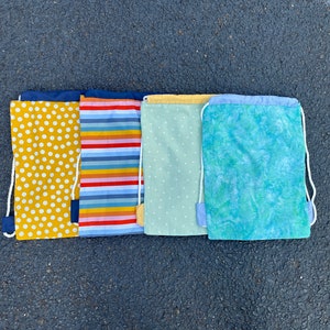 May include: Five drawstring bags with different patterns. The bags are lined up from left to right: yellow with white polka dots, blue and yellow stripes, light green with white polka dots, and light blue with a green and white marbled pattern.