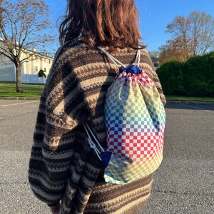 May include: A person wearing a brown and white striped sweater with a rainbow checkered drawstring bag.