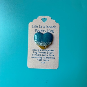 Può includere: Un biglietto bianco con il testo "Life is a beach Pocket Hug" e un ciondolo a forma di cuore in resina blu e oro. Il testo sul biglietto recita: "Here's a little pocket hug for when I can't be there, just a little something to show you that I care XXX."